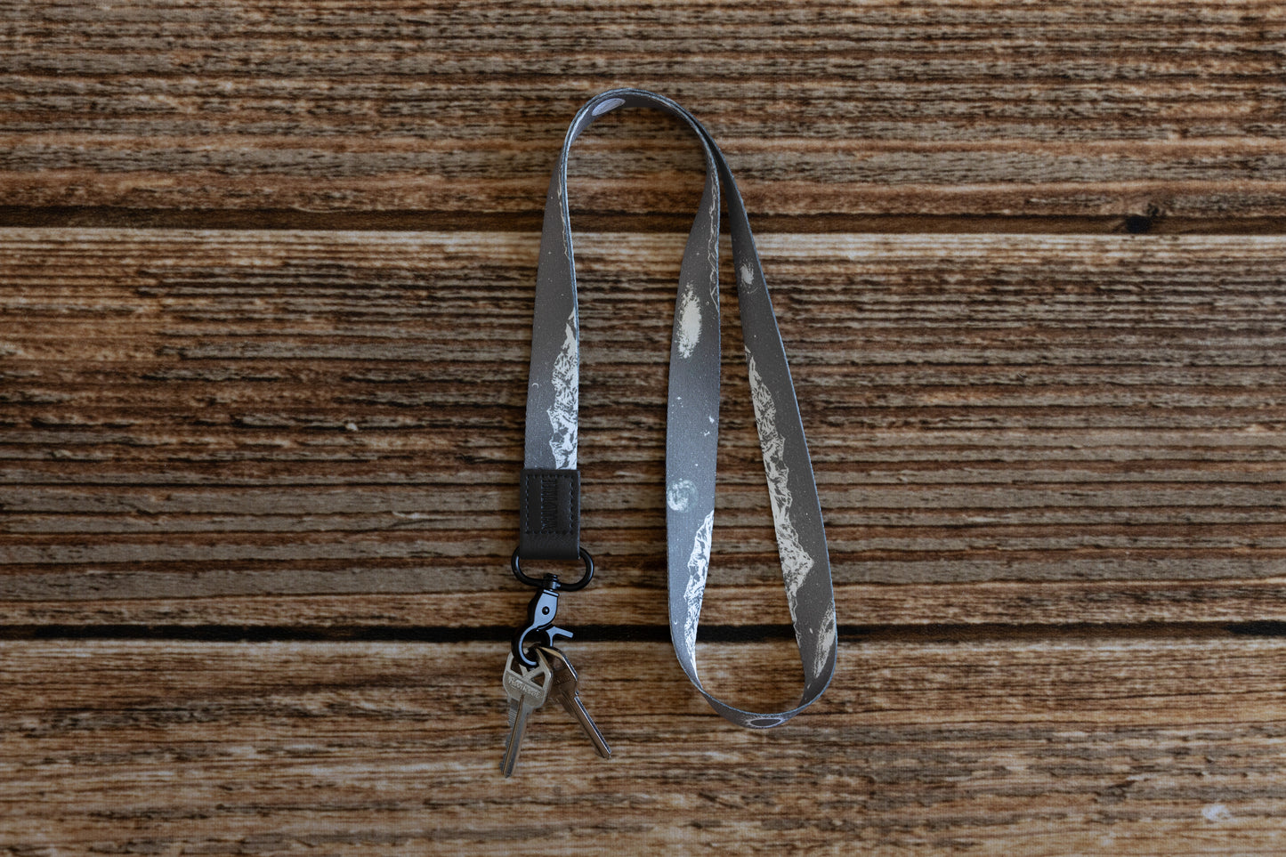 A gray neck lanyard with a white mountain and celestial design, with keys attached to its black metal clasp, lying on a light brown wooden surface.
