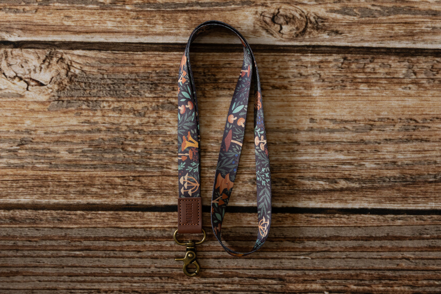 The Forest Foliage Neck Lanyard is arranged in a loop on a textured wooden surface. The lanyard has a dark base with a detailed pattern of different types of mushrooms in orange, brown, and white, complemented by green and blue leaves and berries. A brass-colored clip and a brown faux-leather attachment are visible at the top.