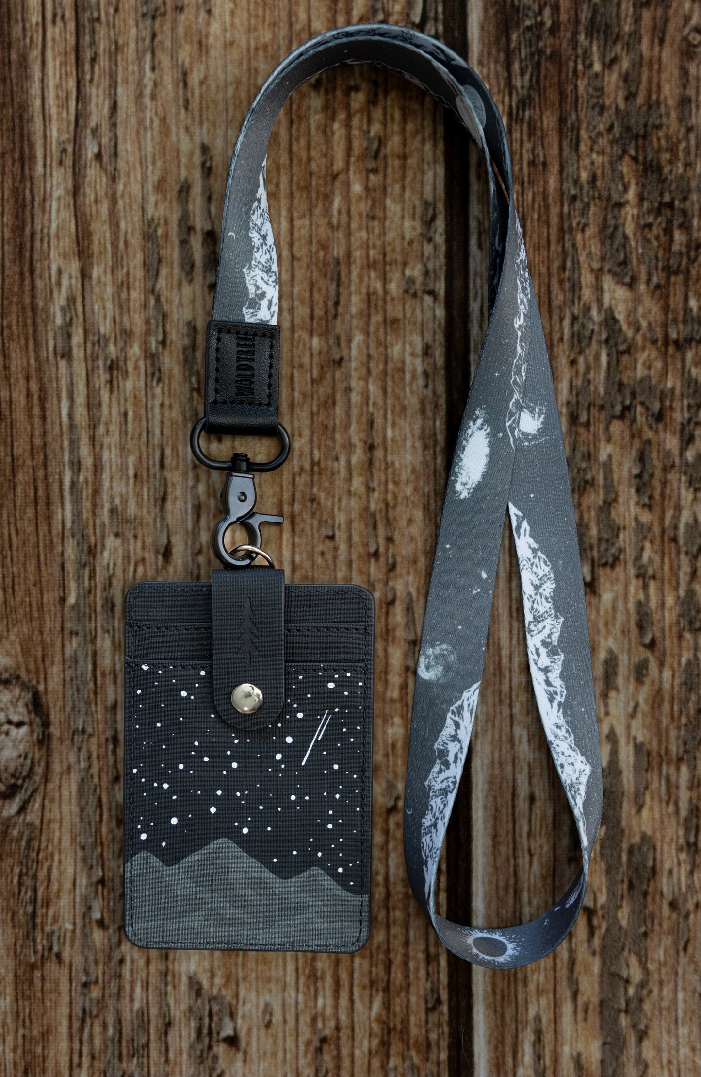 A waist-up lifestyle view of a person outdoors wearing the black and gray Midnight Mountain ID badge holder clipped to a matching mountain-themed neck lanyard. This durable vegan leather vertical badge wallet hangs neatly against a black t-shirt, showing how teachers, nurses, and office workers can wear it comfortably on the job.