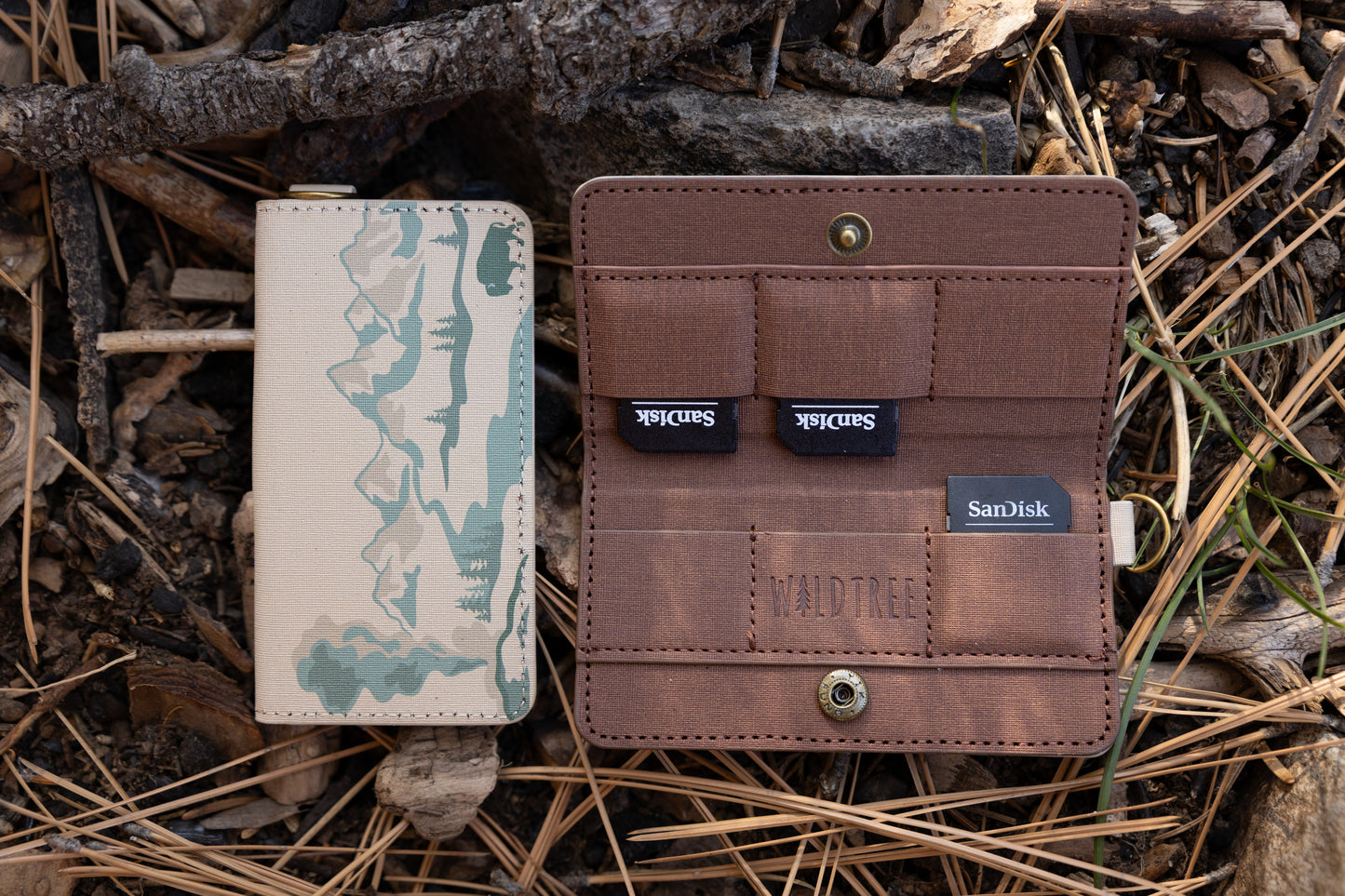 A side-by-side view of a tan fabric SD card holder. The left shows the closed front featuring a teal mountain and forest illustration; the right shows the open interior with brown pockets holding three SanDisk SD cards, featuring the "WILDTREE" logo.