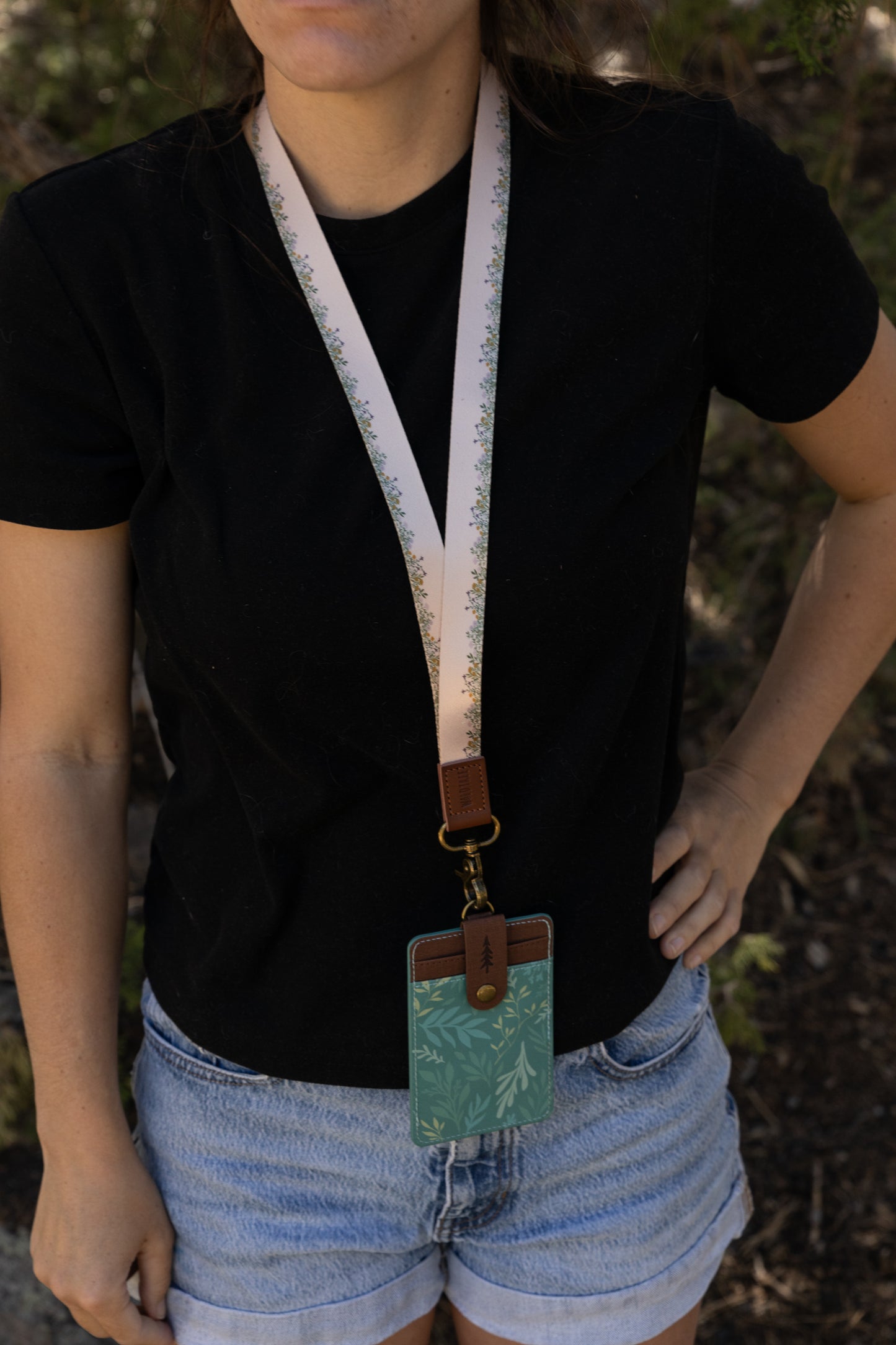 A person wearing a vertical green botanical ID holder clipped to a patterned neck lanyard. The image shows how the product sits comfortably against a black t-shirt when worn outdoors.