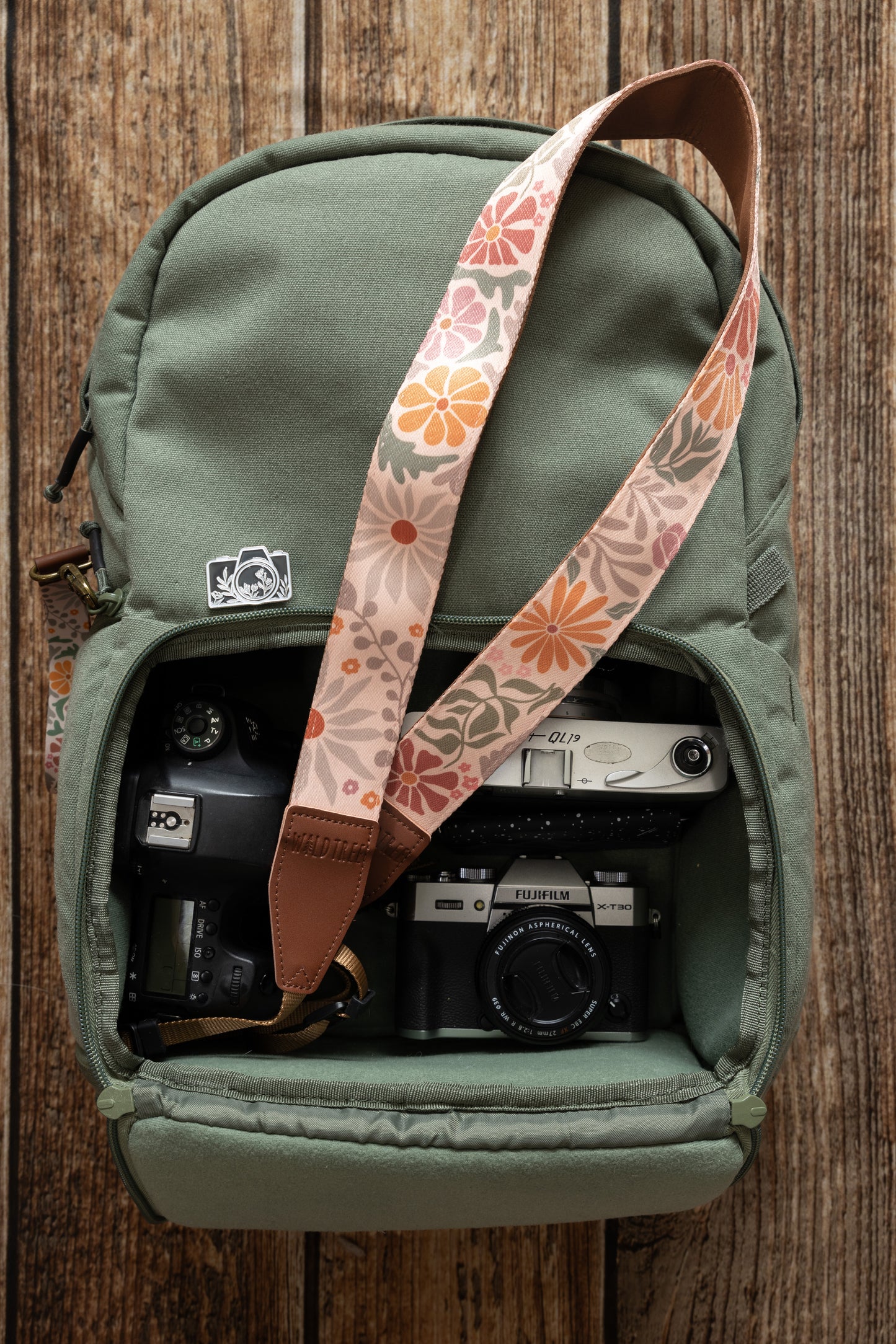 The Sunset Flower camera strap is shown inside an open olive-green camera backpack. The floral pattern and leather-like connectors are visible. Two cameras are also visible inside the backpack.