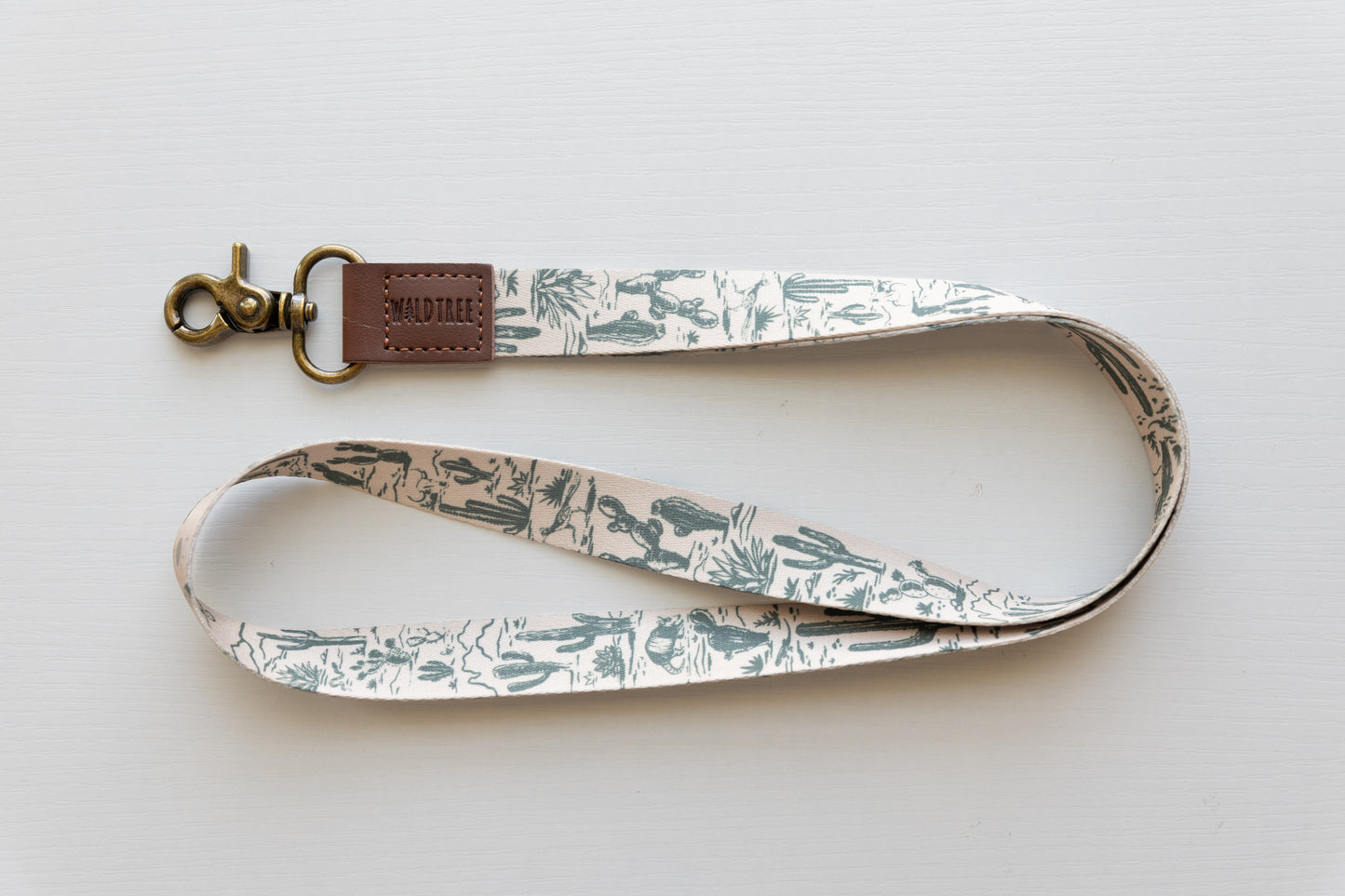 Western landscape neck lanyard by Wildtree, featuring a pattern of green cacti and desert flora on a light background, laid flat on a neutral surface, with a brass-colored swivel clasp and a brown leather tag embossed with "WILDTREE."