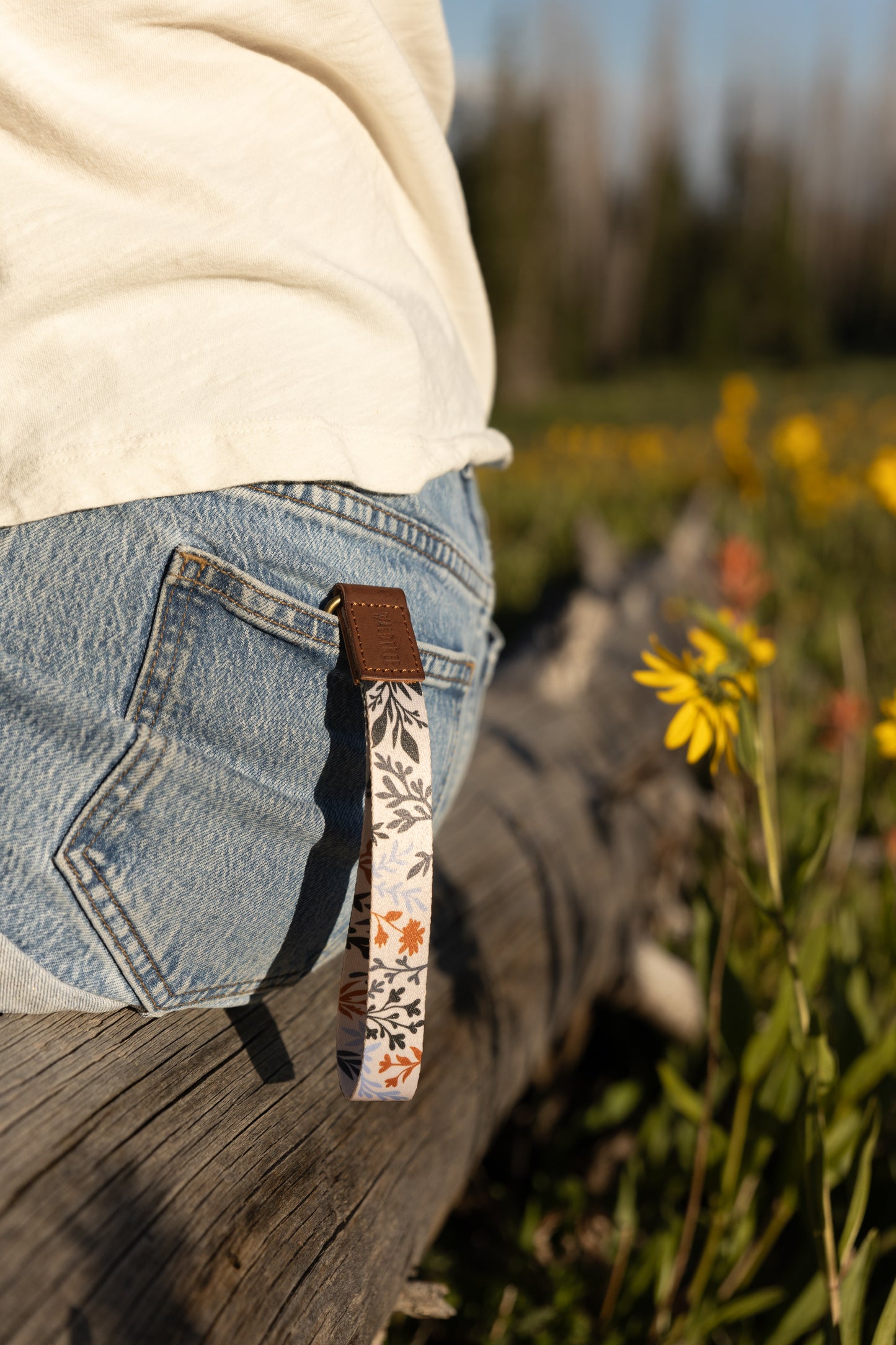 A person sits on a weathered log, and a wristlet keychain with a dark floral and foliage pattern hangs from a belt loop on their denim shorts. A bright yellow wildflower is visible in the foreground to the right.