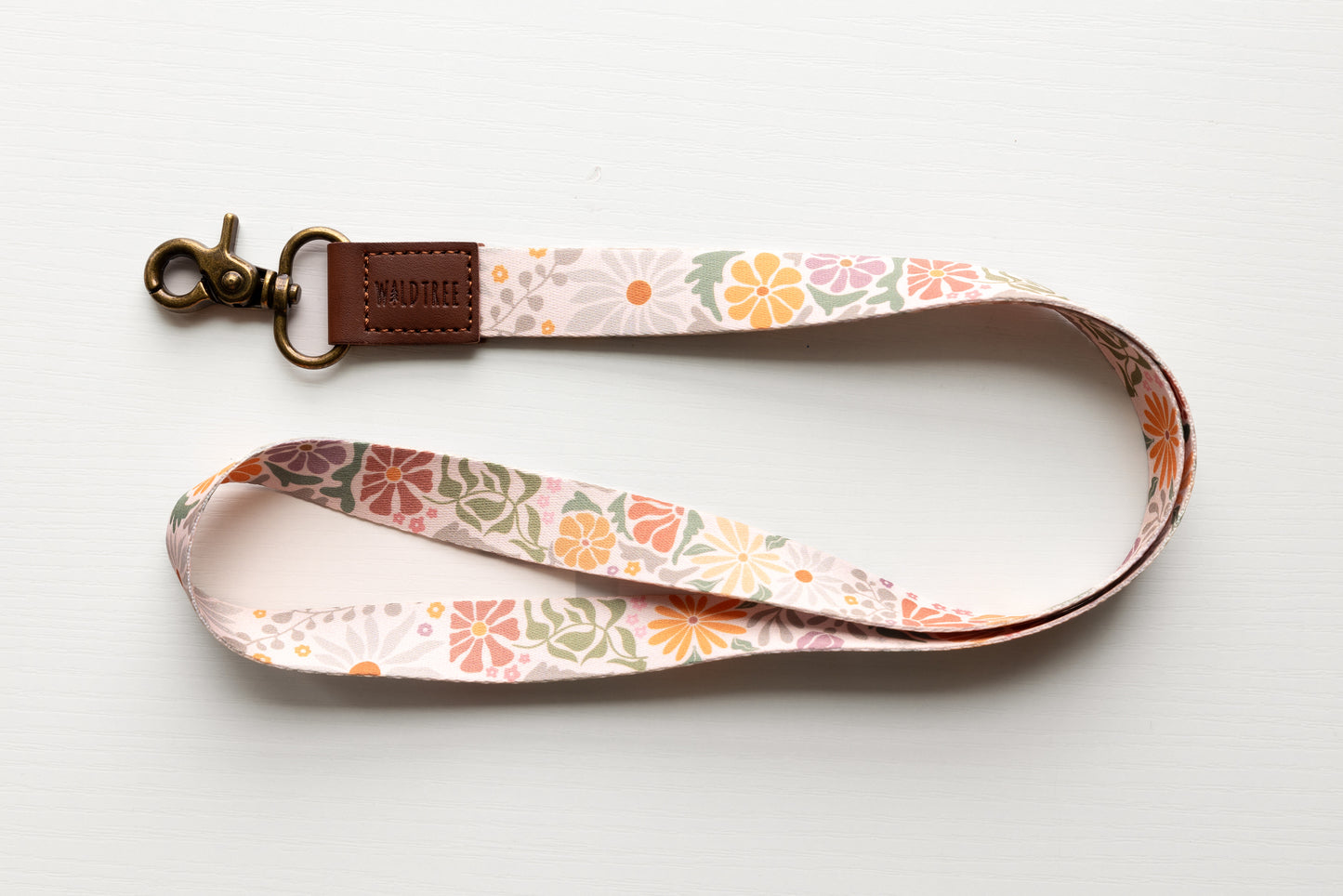 The "Wildtree sunset flower neck lanyard" is showcased against a clean white surface. This stylish lanyard features a light tan fabric strap with vibrant floral designs in orange, pink, and muted green, reminiscent of a warm sunset. A brown leather loop ("WILDTREE") connects to a bronze swivel clasp. The curved lanyard displays the full-length floral print, a fashionable and functional accessory.