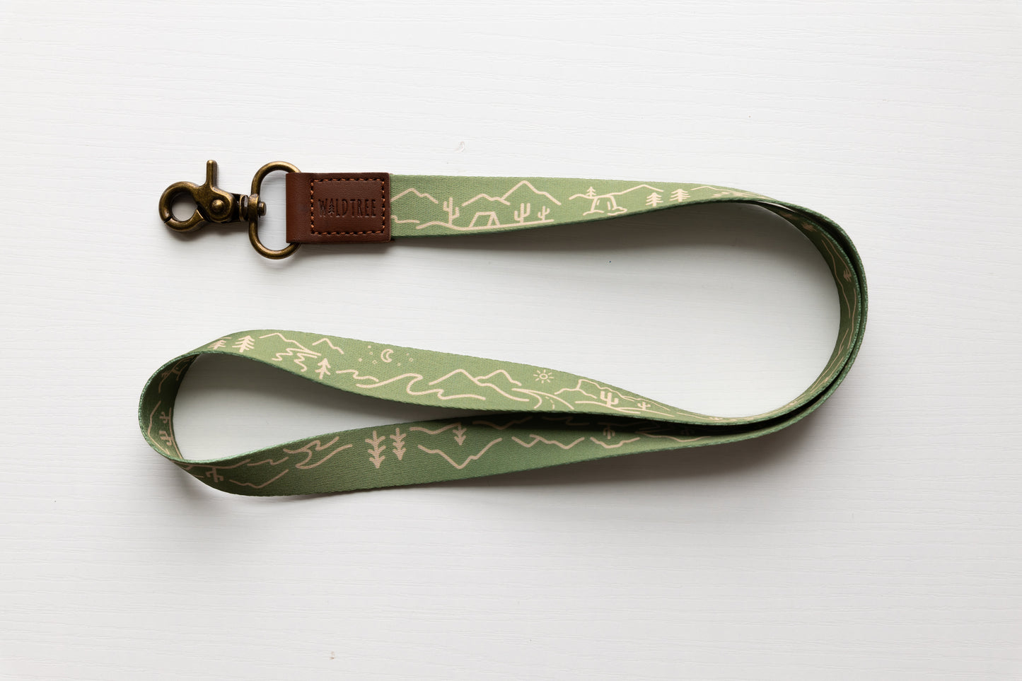 The "Wildtree travel neck lanyard" is presented on a clean light gray surface. The light green strap features a simplistic line art pattern of stylized tents, mountains, cacti, and trees, evoking a sense of wanderlust and outdoor exploration. A brown leather loop ("WILDTREE") securely attaches to a bronze swivel clasp. The gently curved lanyard allows a full view of the continuous travel-inspired artwork.