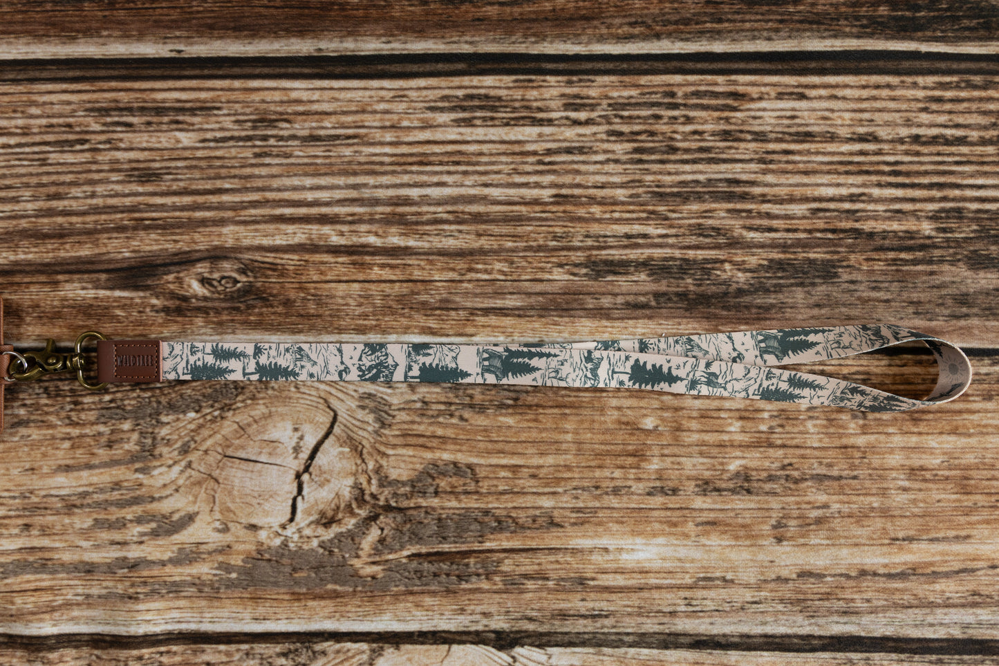 Wildtree Woodland Landscape Neck Lanyard" displayed on a rustic wooden surface. Tan fabric lanyard showcases a nature-inspired print with dark green pine trees and light gray mountain silhouettes. Brown leather loop with "WILDTREE" embossed connects to a bronze-toned metal clasp. Lanyard is presented horizontally, emphasizing the repeating woodland pattern.