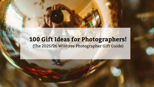 A central white banner with text reading "100 Gift Ideas for Photographers! (The 2025/26 Wildtree Photographer Gift Guide)" is overlaid on a blurry, reflective photo of a person holding a camera, suggesting a holiday ornament or glass surface.