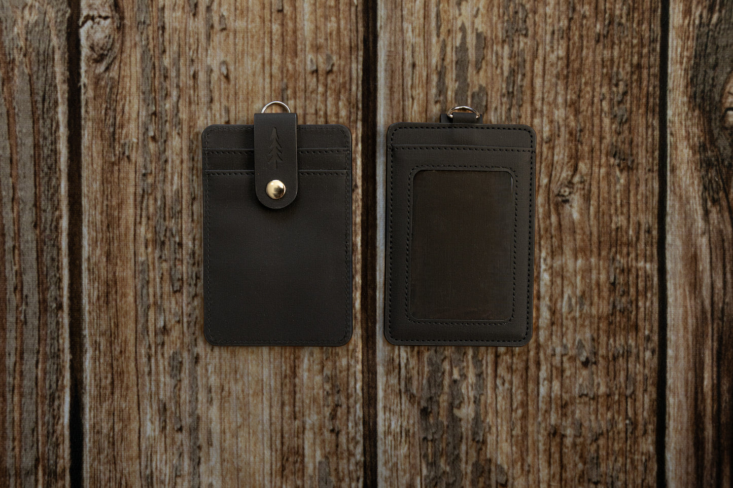 A flat-lay photograph showing the front and back of a dark textured ID badge holder, placed side-by-side on a rustic, weathered wooden surface. The front of the holder features two card slots secured by a vertical strap with an embossed pine tree logo and a brass snap button. The back features a clear plastic window for displaying an ID card.