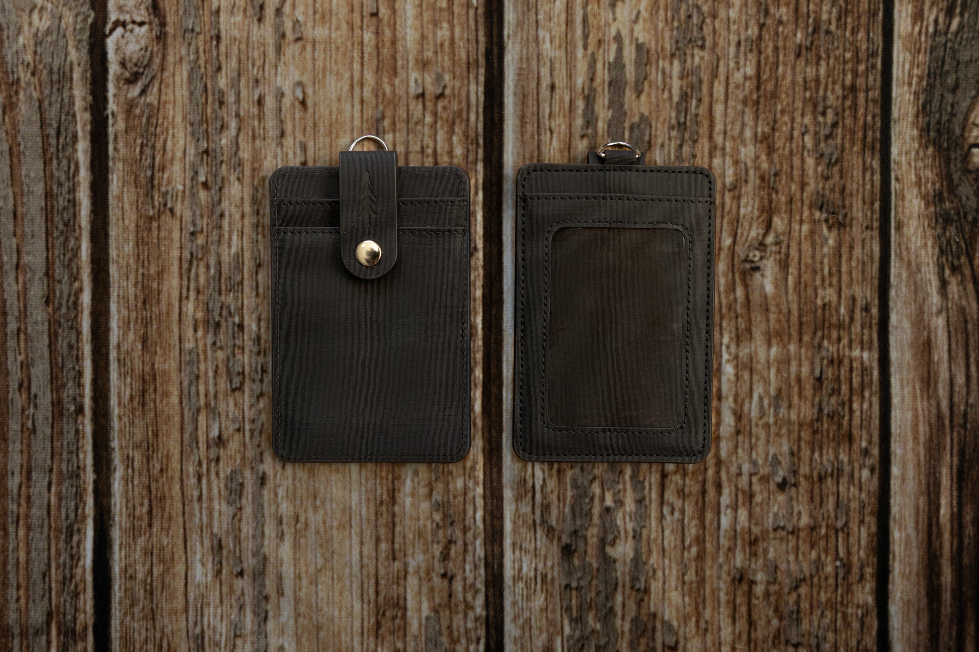 A flat-lay photograph showing the front and back of a dark textured ID badge holder, placed side-by-side on a rustic, weathered wooden surface. The front of the holder features two card slots secured by a vertical strap with an embossed pine tree logo and a brass snap button. The back features a clear plastic window for displaying an ID card.