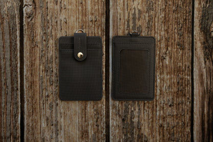 A flat-lay photograph showing the front and back of a dark textured ID badge holder, placed side-by-side on a rustic, weathered wooden surface. The front of the holder features two card slots secured by a vertical strap with an embossed pine tree logo and a brass snap button. The back features a clear plastic window for displaying an ID card.