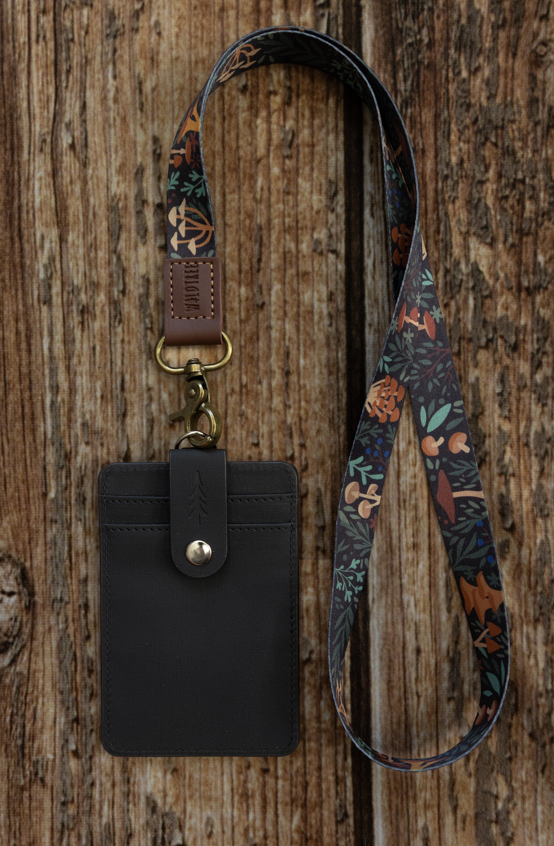 A flat-lay photograph on a vertical wooden plank background showing the front side of the dark textured ID badge holder attached to a different lanyard. This lanyard features a colorful forest floor pattern with various mushrooms, leaves, and ferns on a dark background. It connects to the holder with an antique brass-colored lobster claw clasp and a brown leather loop embossed with "WILDTREE".