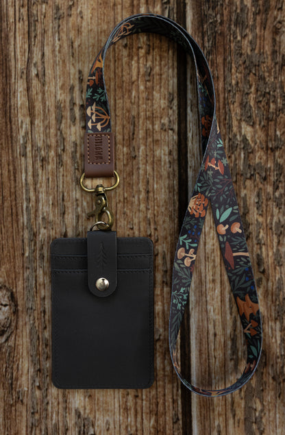 A flat-lay photograph on a vertical wooden plank background showing the front side of the dark textured ID badge holder attached to a different lanyard. This lanyard features a colorful forest floor pattern with various mushrooms, leaves, and ferns on a dark background. It connects to the holder with an antique brass-colored lobster claw clasp and a brown leather loop embossed with "WILDTREE".
