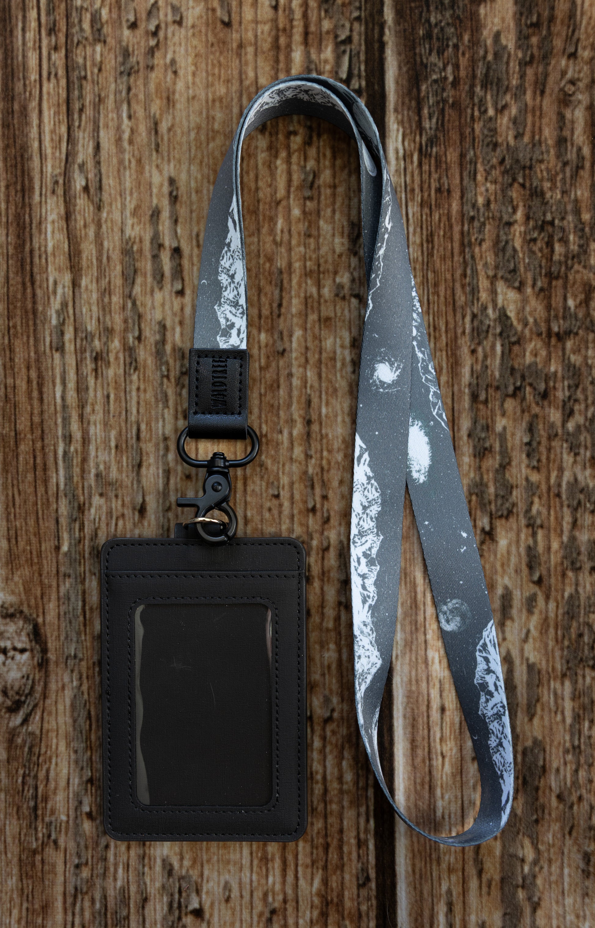 A flat-lay photograph on a vertical wooden plank background showing the back side of the dark textured ID badge holder attached to the same monochrome mountain and galaxy lanyard. The clear plastic ID window is visible on this side, and the lanyard is attached via the black lobster claw clasp.