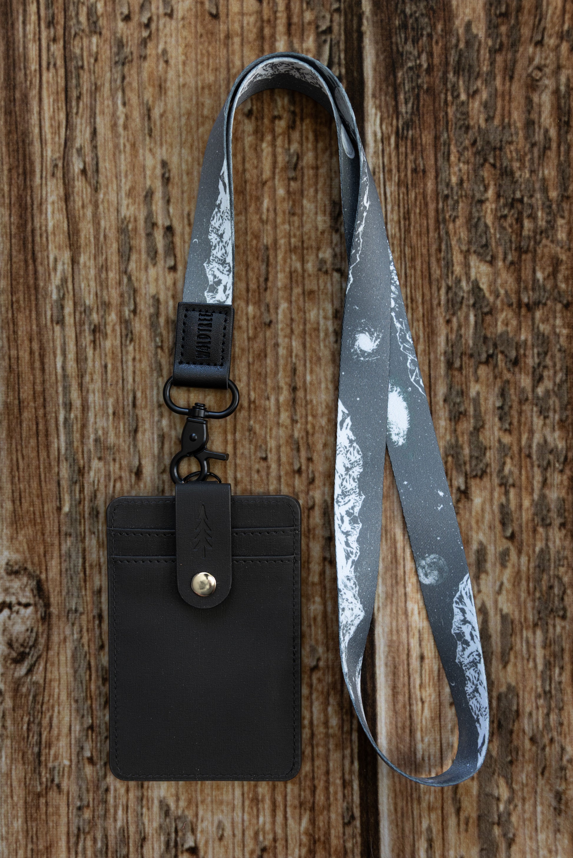 A flat-lay photograph on a vertical wooden plank background showing the front side of a dark textured ID badge holder attached to a patterned lanyard. The lanyard features a monochrome gray-and-white print of mountain peaks, stars, and galaxies. It connects to the badge holder's top D-ring using a matte black lobster claw clasp and a black leather loop embossed with the word "WILDTREE".