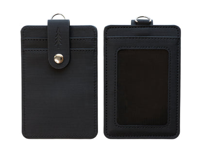 A clean, overhead studio photograph showing the front and back of the dark textured ID badge holder positioned side-by-side against a plain, bright white background. The fine grid texture of the material, the stitching details, and the embossed pine tree on the snap closure strap are clearly visible.