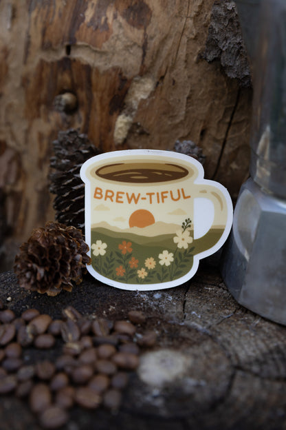 A close-up lifestyle photo featuring a "Brew-tiful" retro campfire mug sticker  centered on a rustic wood tree slice. The background is a detailed weathered tree bark texture. In the foreground, out-of-focus roasted coffee beans and several pinecones are artfully scattered, with a silver Moka pot coffee maker visible on the right.