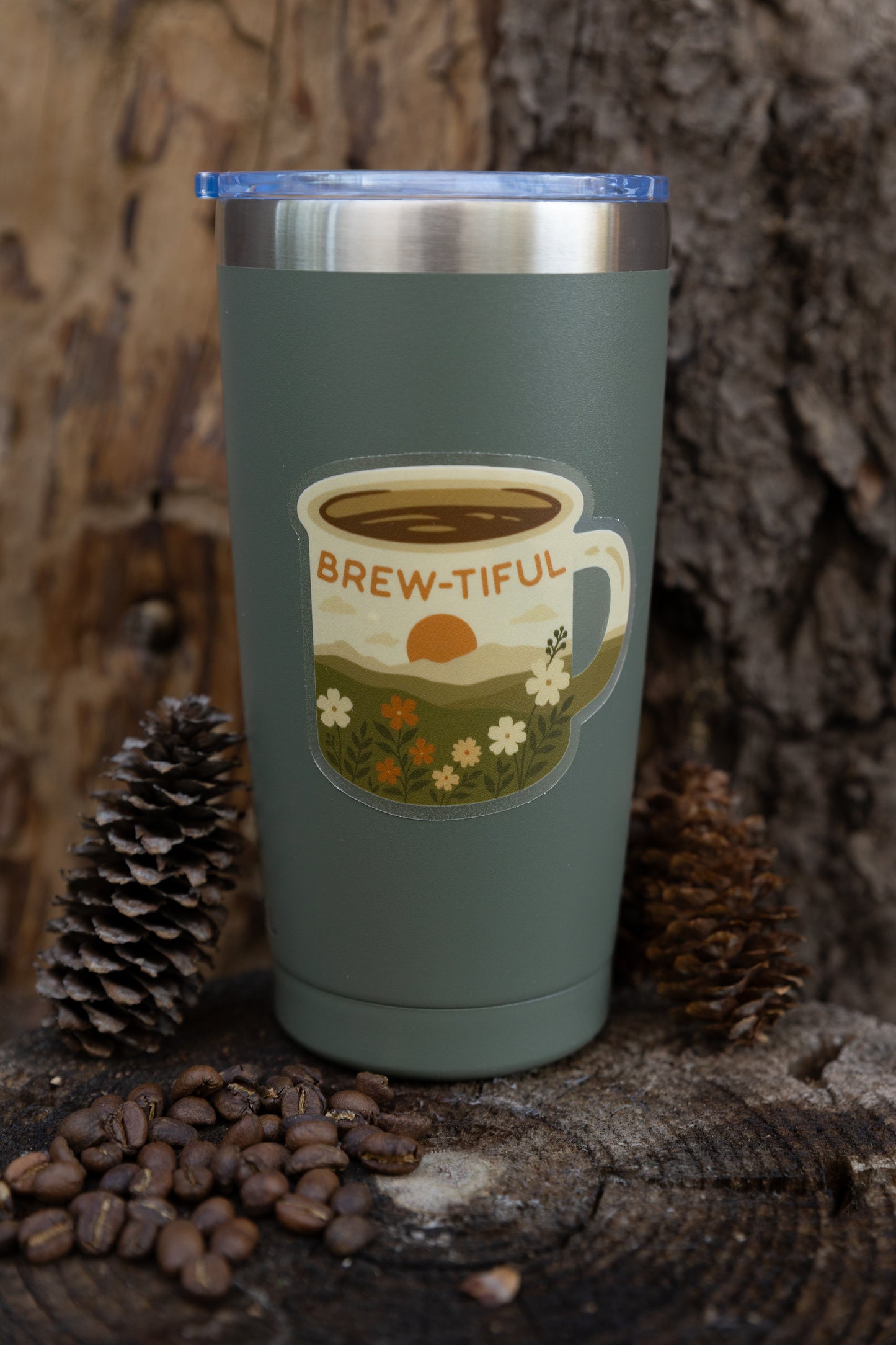 A product showcase photo displaying the "Brew-tiful" retro coffee mug sticker applied to the center of a matte, muted sage green stainless steel travel tumbler with a clear lid. The tumbler rests on a weathered wood slice background, flanked by two large pinecones and a pile of whole roasted coffee beans, highlighting the sticker's suitability for water bottles.
