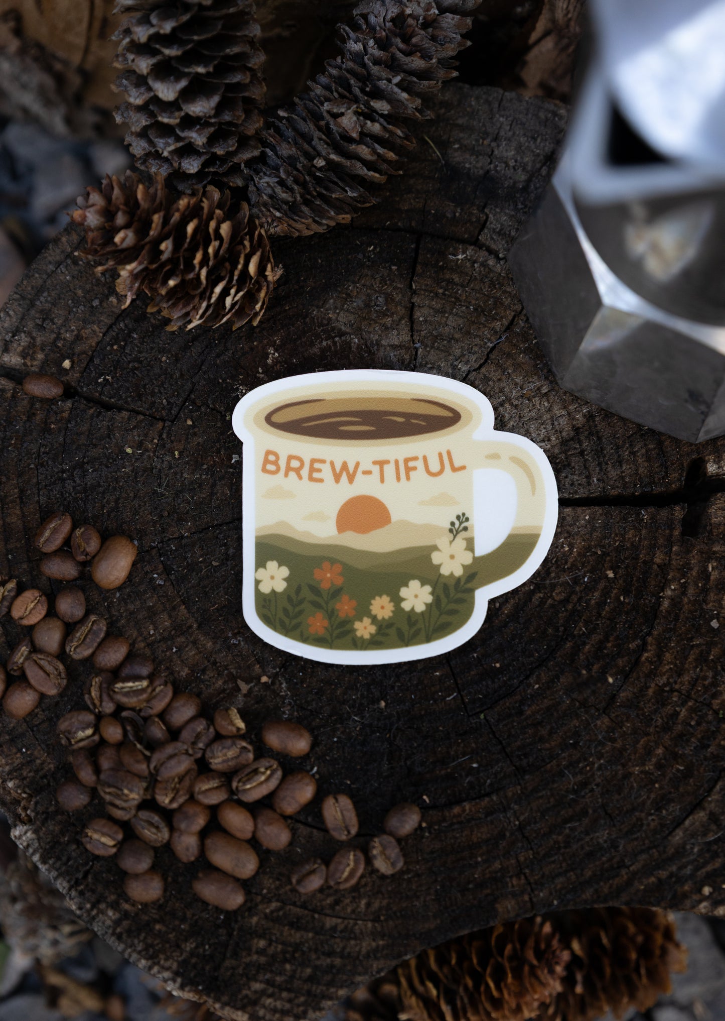 An overhead flat lay photograph centered on the "Brew-tiful" die-cut vinyl sticker. The sticker, which features a retro mountain sunset and wildflower meadow design, rests directly on the circular surface of a dark wood tree stump. Whole roasted coffee beans are scattered in the lower left corner, with two pinecones and a portion of a silver Moka pot framing the top and bottom.