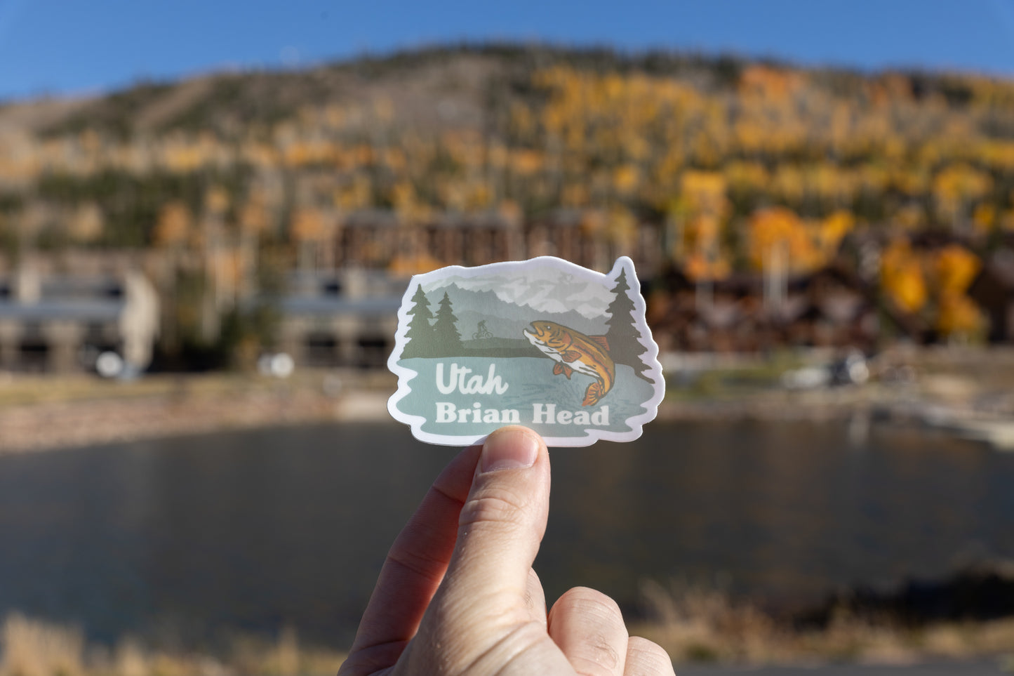 A hand holds the Brian Head, Utah sticker featuring a jumping trout, mountains, and pine trees. The sticker is held over a dark, calm body of water (likely a pond). In the blurry background, a hillside is covered in bright yellow and orange autumn foliage with several rustic mountain buildings visible at the base of the hill.