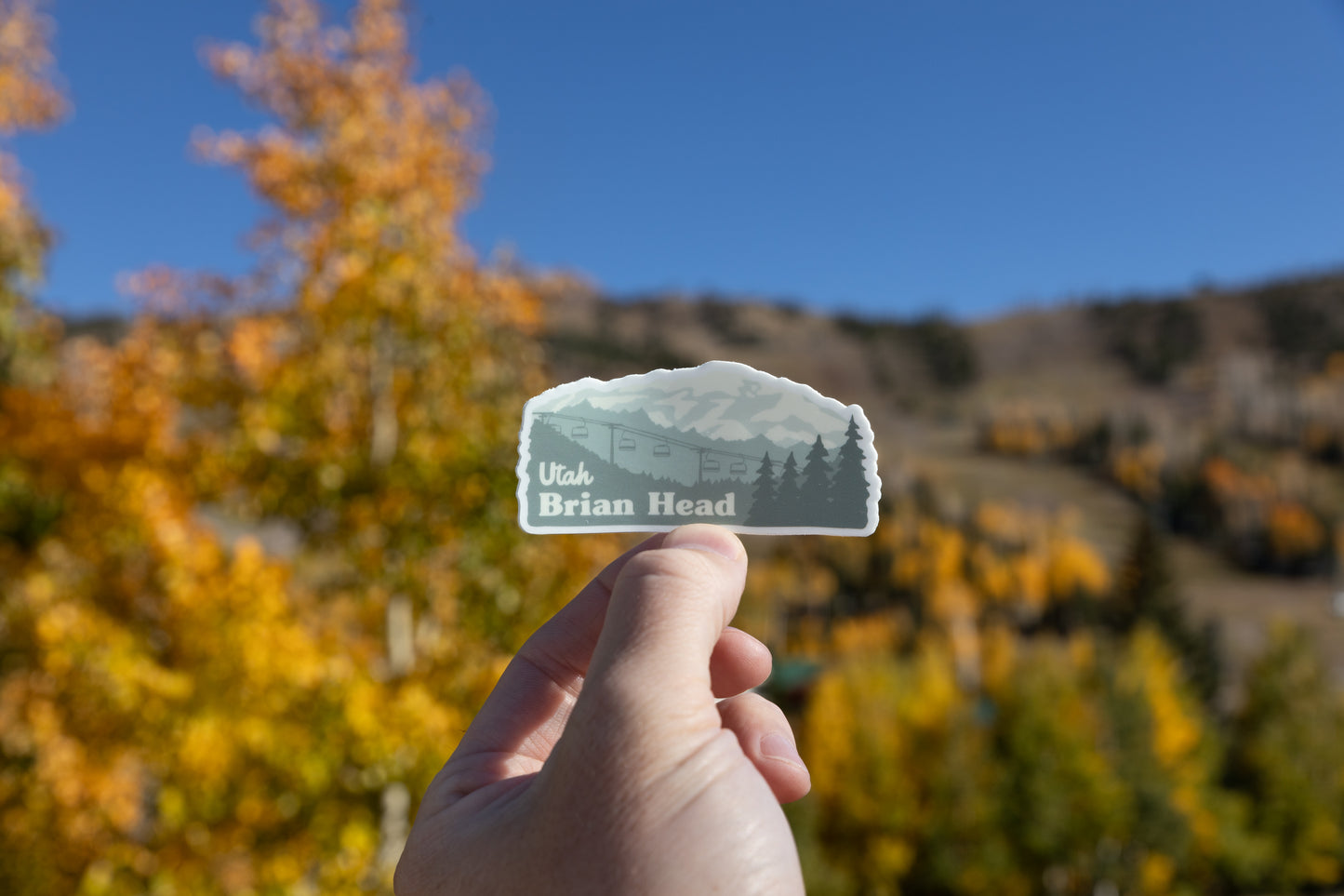 A hand holds up a Brian Head, Utah sticker outdoors. The sticker, which shows a mountain, a ski lift, and pine trees, is held against a vibrant background of blurry yellow and orange autumn foliage beneath a clear blue sky, suggesting the photograph was taken on the mountain during the fall season.
