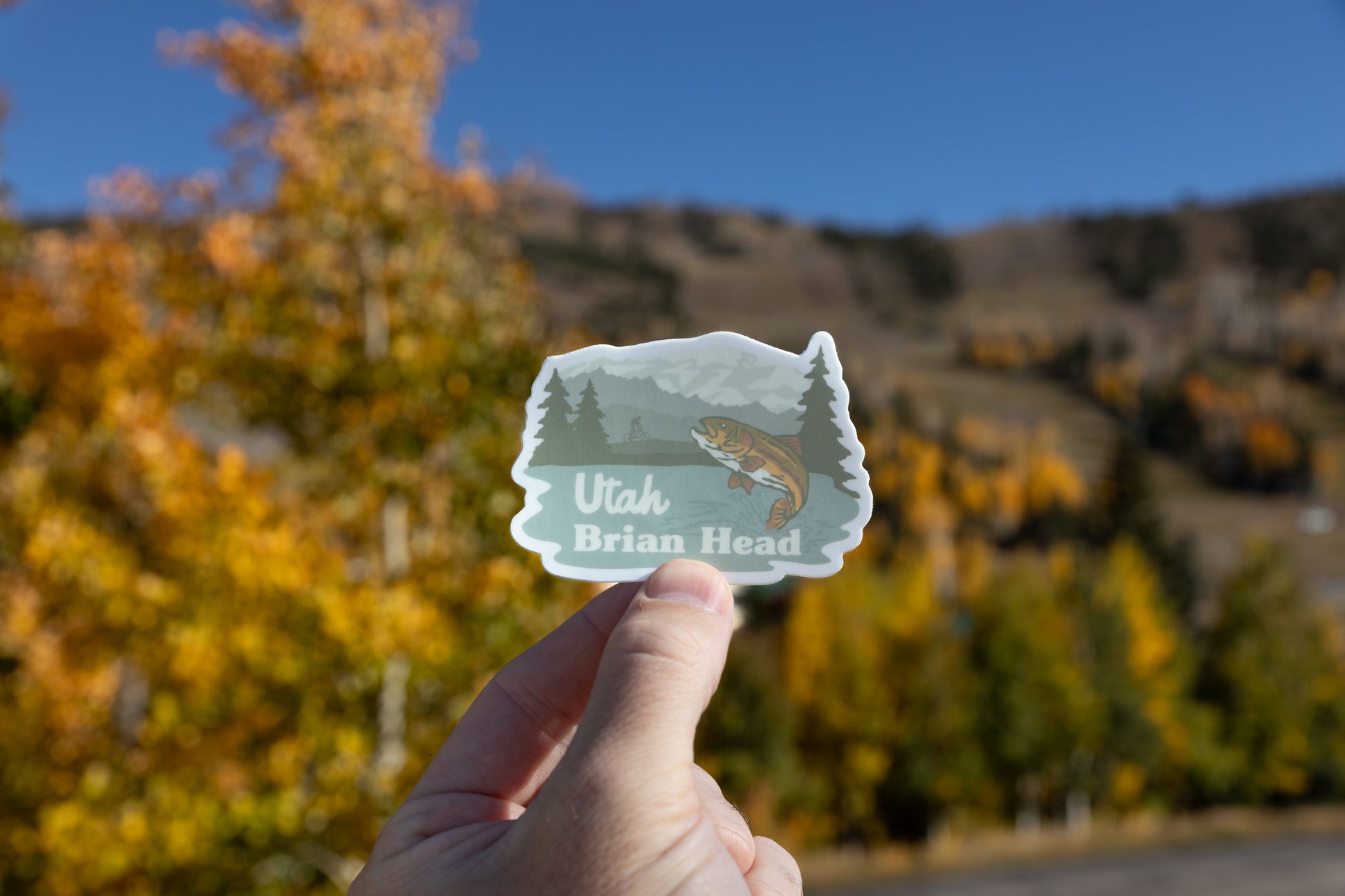 A hand holds up the Brian Head, Utah outdoor recreation sticker against a beautiful mountain background. The sticker illustration shows a trout jumping and a distant cyclist. The background is brightly lit by the sun, featuring a clear blue sky above a hillside covered in blurry yellow and orange autumn aspen trees.