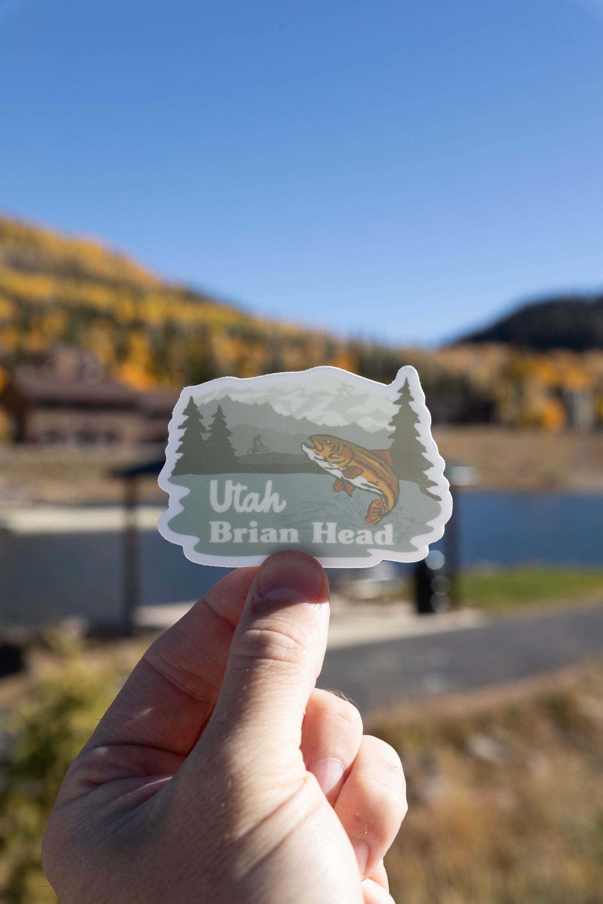 A vertical photograph shows a hand holding the Brian Head, Utah sticker featuring a jumping trout, mountains, and a cyclist. The sticker is held against an outdoor background that includes a dark blue body of water (likely a pond) and a blur of yellow-leaved trees on the far hill under a clear, bright blue sky.