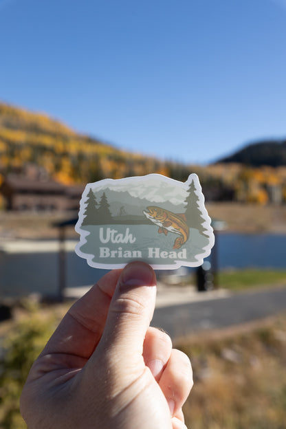 A vertical photograph shows a hand holding the Brian Head, Utah sticker featuring a jumping trout, mountains, and a cyclist. The sticker is held against an outdoor background that includes a dark blue body of water (likely a pond) and a blur of yellow-leaved trees on the far hill under a clear, bright blue sky.