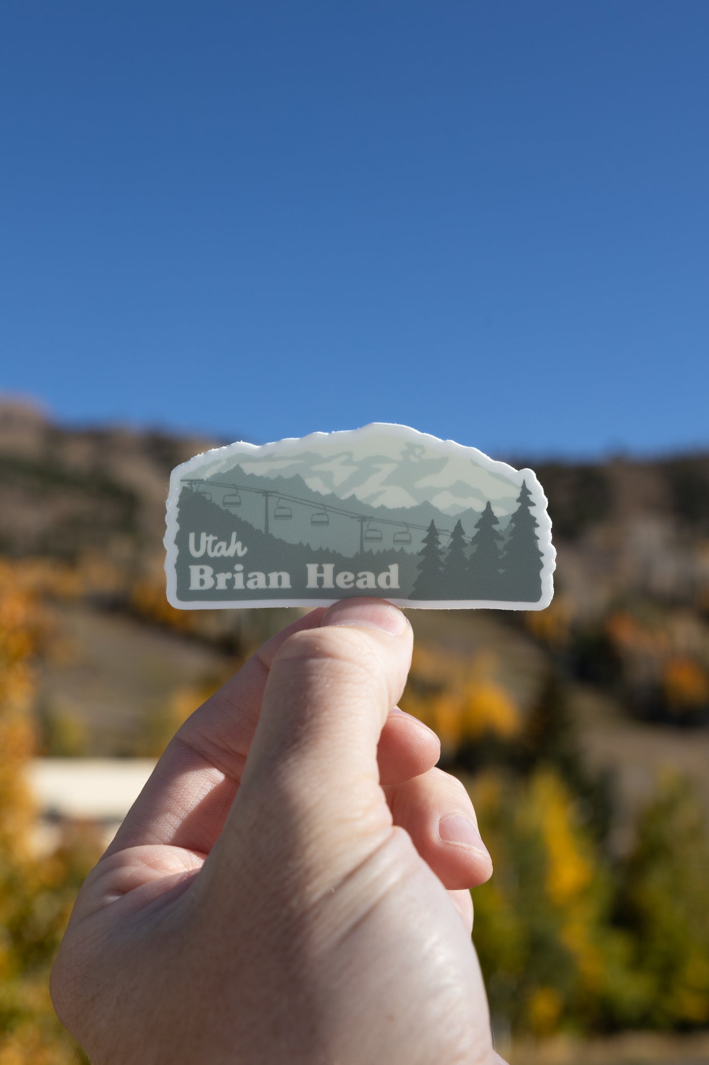 A vertical photo shows a hand holding a Brian Head, Utah sticker in the foreground. The sticker design features a mountain, ski lift, and trees. The background is a clear, vibrant blue sky above a blurry mountain slope, with hints of yellow and brown autumn foliage visible below.