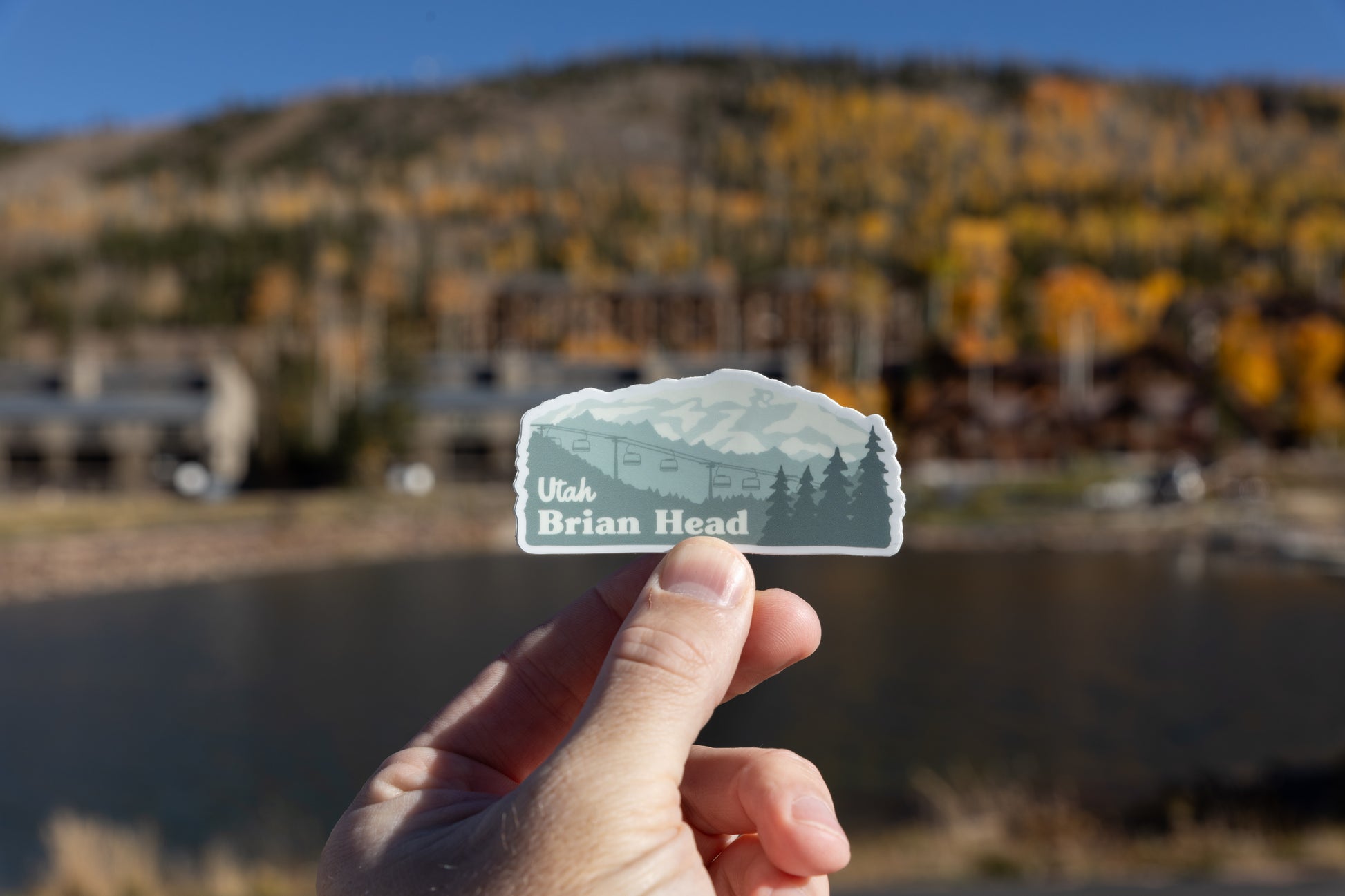 close-up photograph of a hand holding the Brian Head, Utah sticker in the foreground. The sticker is positioned over a dark body of water (likely a pond or small lake) in the midground. The background is a blurry hillside covered in autumn trees with yellow foliage and several dark, rustic buildings or lodges on the far shore.