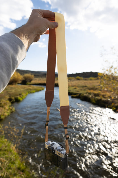 Honey yellow camera strap with brown leather ends connected to fujifilm camera being held up by hand in front of river and valley