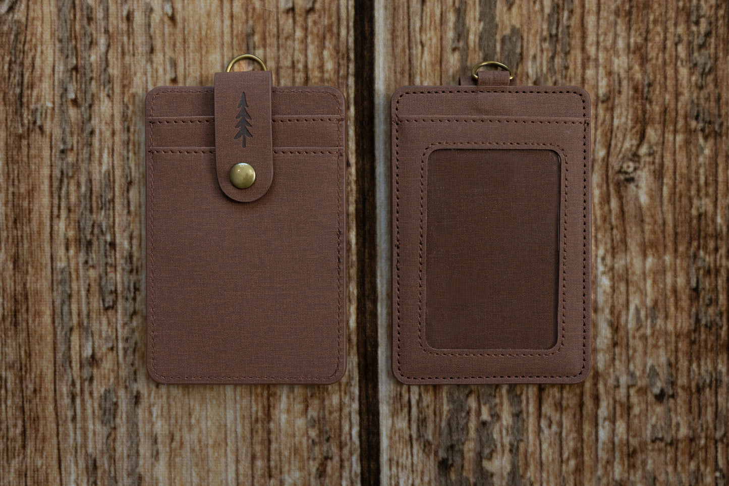 Two brown textured ID badge holders shown side-by-side against a rustic wooden backdrop. The left holder displays the front side, featuring card slots and a snap-button closure strap embossed with a pine tree icon. The right holder displays the back side, featuring a clear plastic window for displaying an ID card.