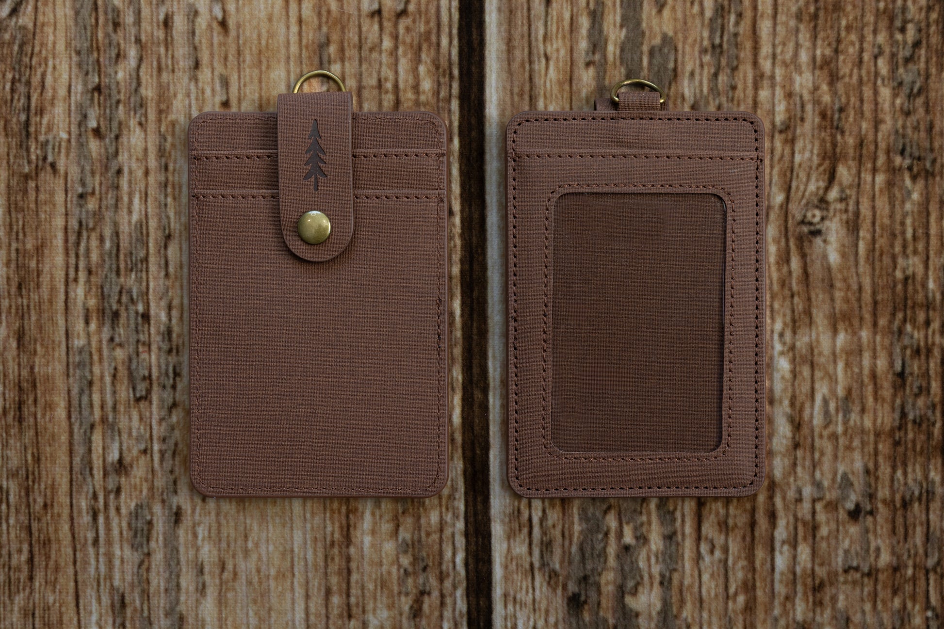 Two brown textured ID badge holders shown side-by-side against a rustic wooden backdrop. The left holder displays the front side, featuring card slots and a snap-button closure strap embossed with a pine tree icon. The right holder displays the back side, featuring a clear plastic window for displaying an ID card.