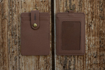 Two brown textured ID badge holders shown side-by-side against a rustic wooden backdrop. The left holder displays the front side, featuring card slots and a snap-button closure strap embossed with a pine tree icon. The right holder displays the back side, featuring a clear plastic window for displaying an ID card.