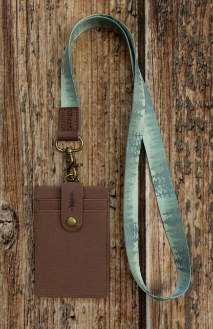 A brown textured ID badge holder attached to a light sage green fabric lanyard. The lanyard displays a subtle, repeating forest silhouette pattern of pine trees in varying shades of green. The assembly is photographed against a rustic wooden background.