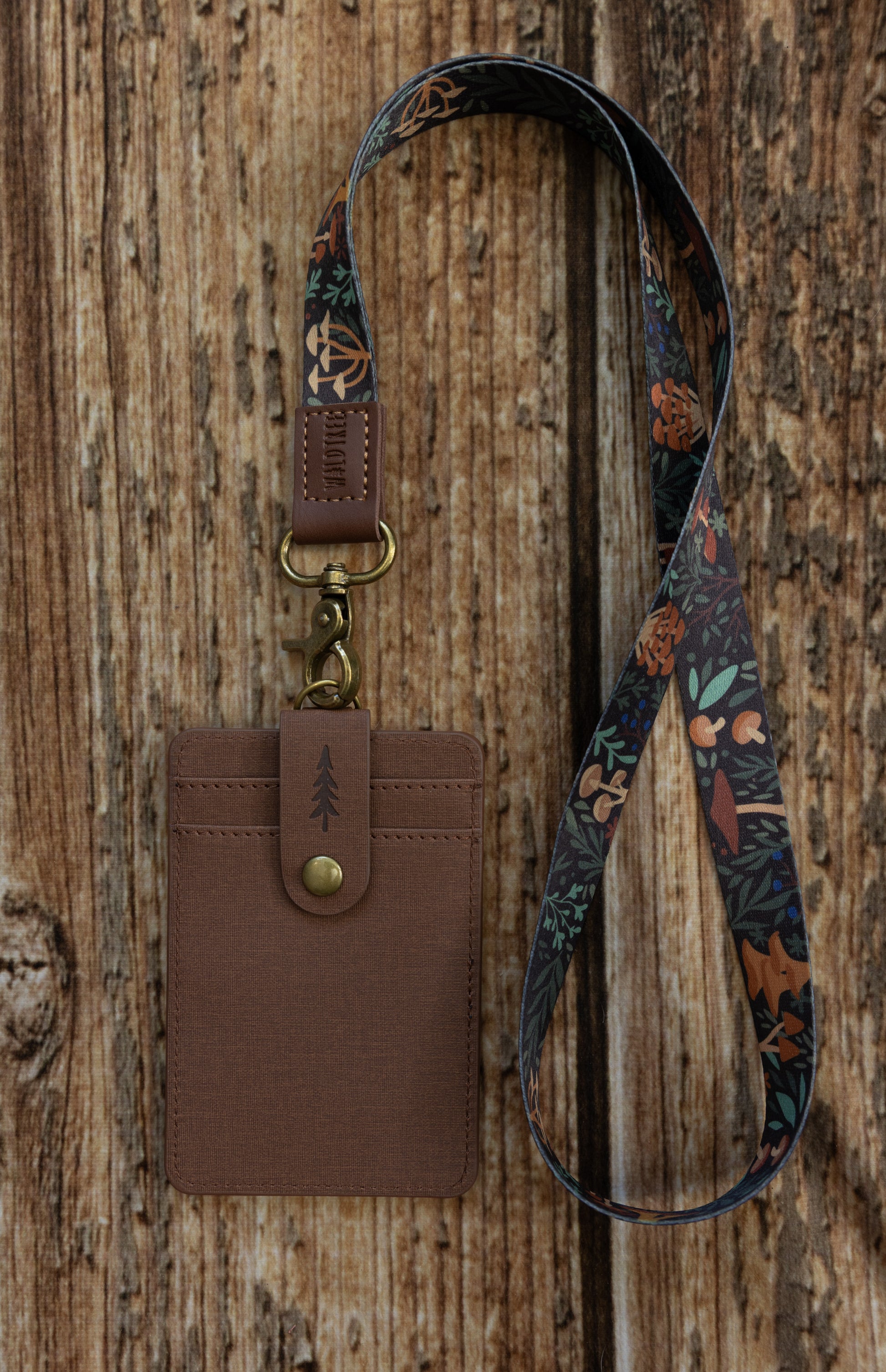 A brown textured ID badge holder attached to a dark, multi-colored fabric lanyard. The lanyard is decorated with an illustrated pattern of various mushrooms, ferns, and forest foliage in shades of orange, green, and brown. The item is displayed vertically against a rustic wooden backdrop.