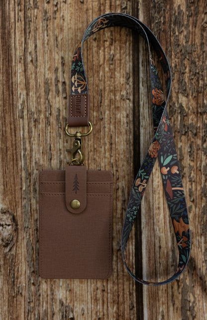 A brown textured ID badge holder attached to a dark, multi-colored fabric lanyard. The lanyard is decorated with an illustrated pattern of various mushrooms, ferns, and forest foliage in shades of orange, green, and brown. The item is displayed vertically against a rustic wooden backdrop.