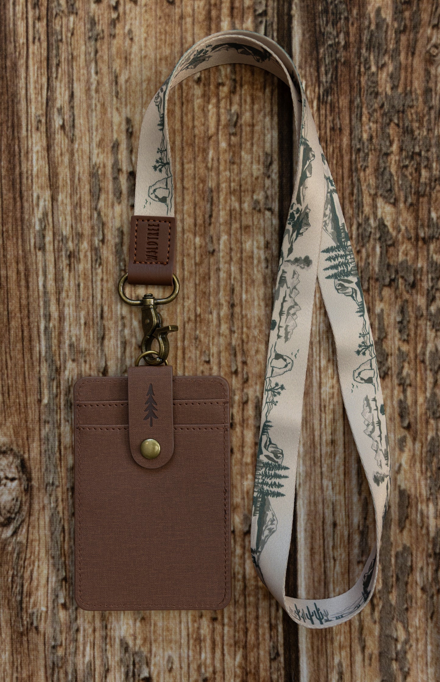 A brown textured ID badge holder attached to a light beige fabric national park lanyard. The lanyard features a dark green illustrated pattern of desert landscapes, mountains, rock arches, cacti, and pine trees. The badge holder has a snap closure strap embossed with a pine tree icon and is set against a rustic wooden background.