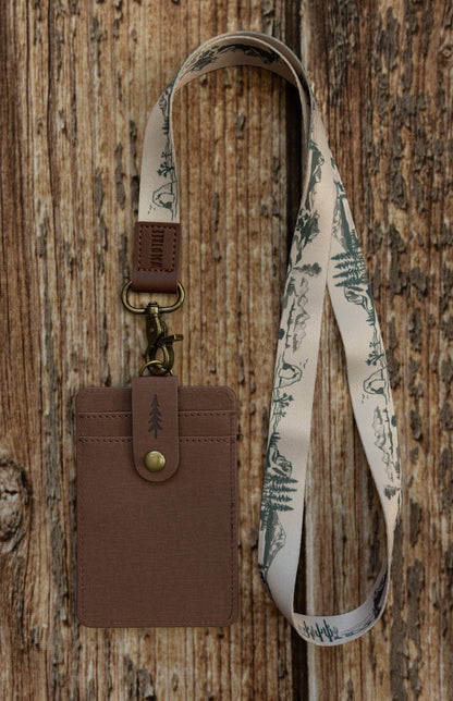 A brown textured ID badge holder attached to a light beige fabric national park lanyard. The lanyard features a dark green illustrated pattern of desert landscapes, mountains, rock arches, cacti, and pine trees. The badge holder has a snap closure strap embossed with a pine tree icon and is set against a rustic wooden background.