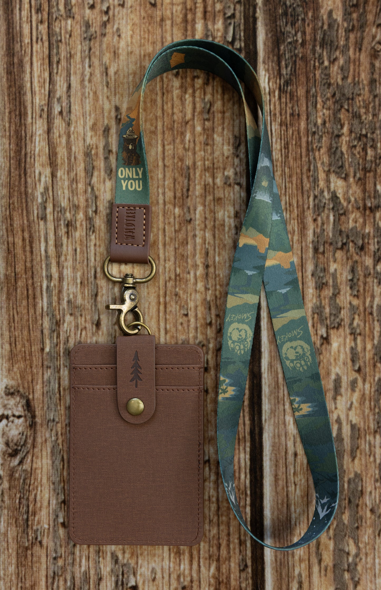 A brown textured ID badge holder clipped to a dark green and camouflage-style fabric lanyard. The lanyard features a Smokey Bear theme, showing illustrations of Smokey Bear wearing his campaign hat, his paw print, and the phrase "ONLY YOU." The product rests vertically against a rustic wooden surface.
