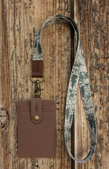A brown textured ID badge holder connected to a light beige fabric lanyard. The lanyard features a detailed dark green sketchy illustration of a forest scene with pine trees and a bear walking through the woods. The badge holder is suspended against a textured wood panel background.