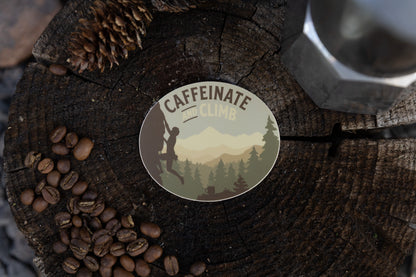 A high-angle close-up of the "Caffeinate and Climb" climbing decal positioned on a rustic wood surface. To the left, a pile of coffee beans is scattered near two pinecones, with a silver Moka pot partially visible in the blurred background.