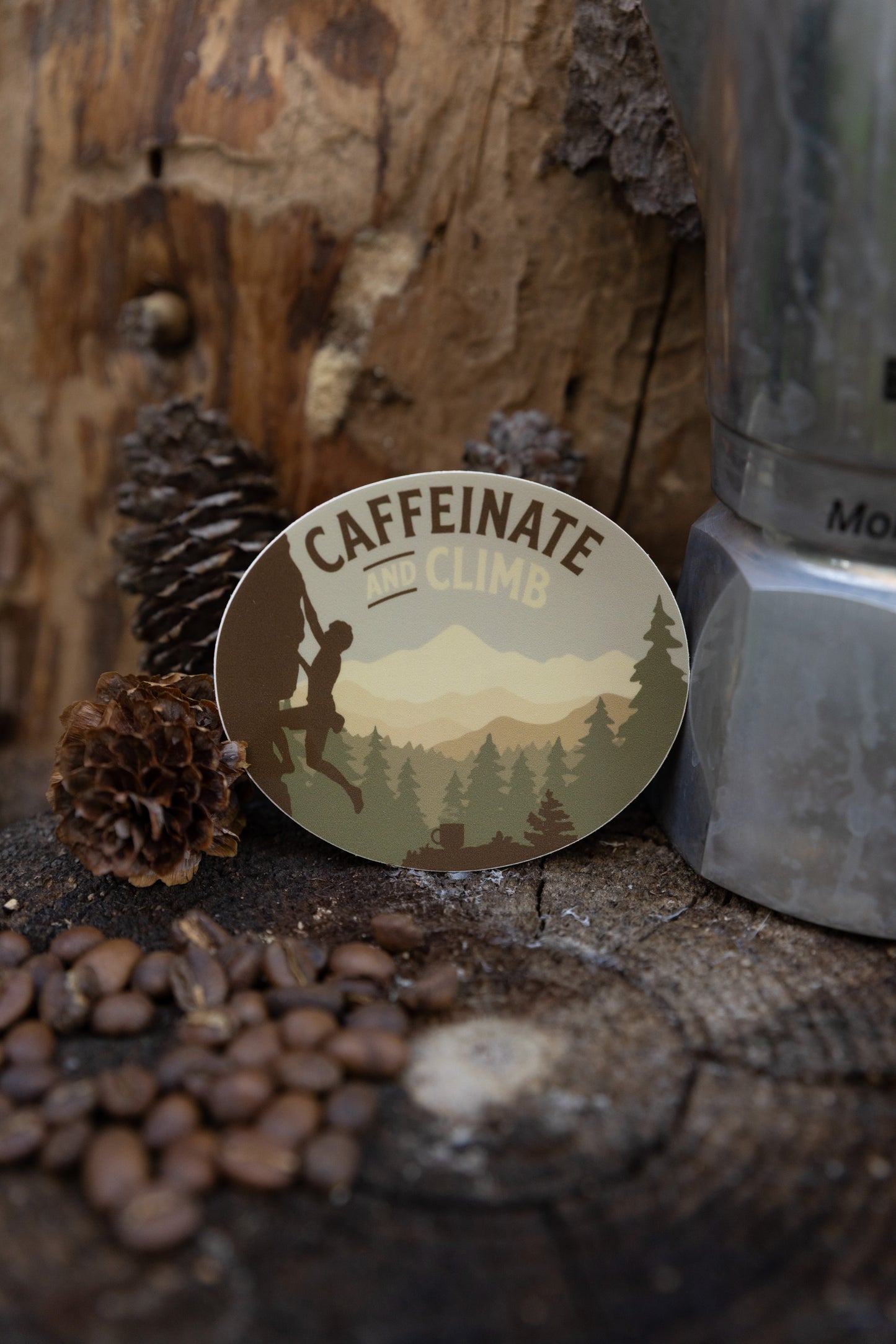 A lifestyle shot of the "Caffeinate and Climb" sticker standing against a piece of rough tree bark. The foreground features scattered coffee beans and pinecones on a wood stump, creating a rugged, outdoorsy atmosphere next to a silver coffee pot.