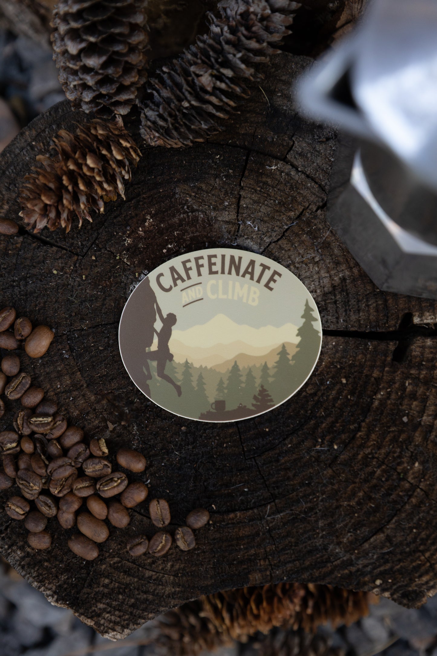 An overhead flat lay of the circular "Caffeinate and Climb" sticker resting on a dark, weathered wood tree stump. The scene is framed by roasted coffee beans, dried pinecones, and the edge of a silver metal coffee maker.