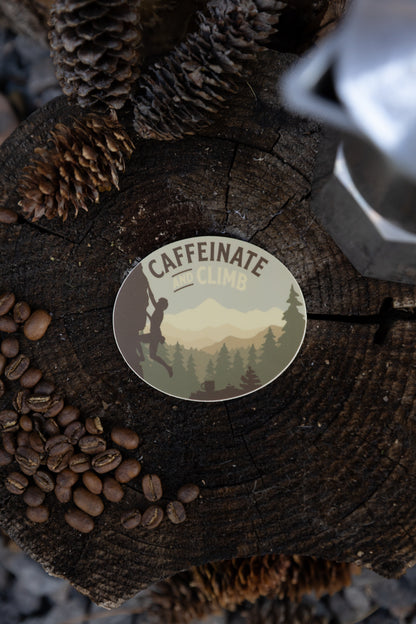 An overhead flat lay of the circular "Caffeinate and Climb" sticker resting on a dark, weathered wood tree stump. The scene is framed by roasted coffee beans, dried pinecones, and the edge of a silver metal coffee maker.