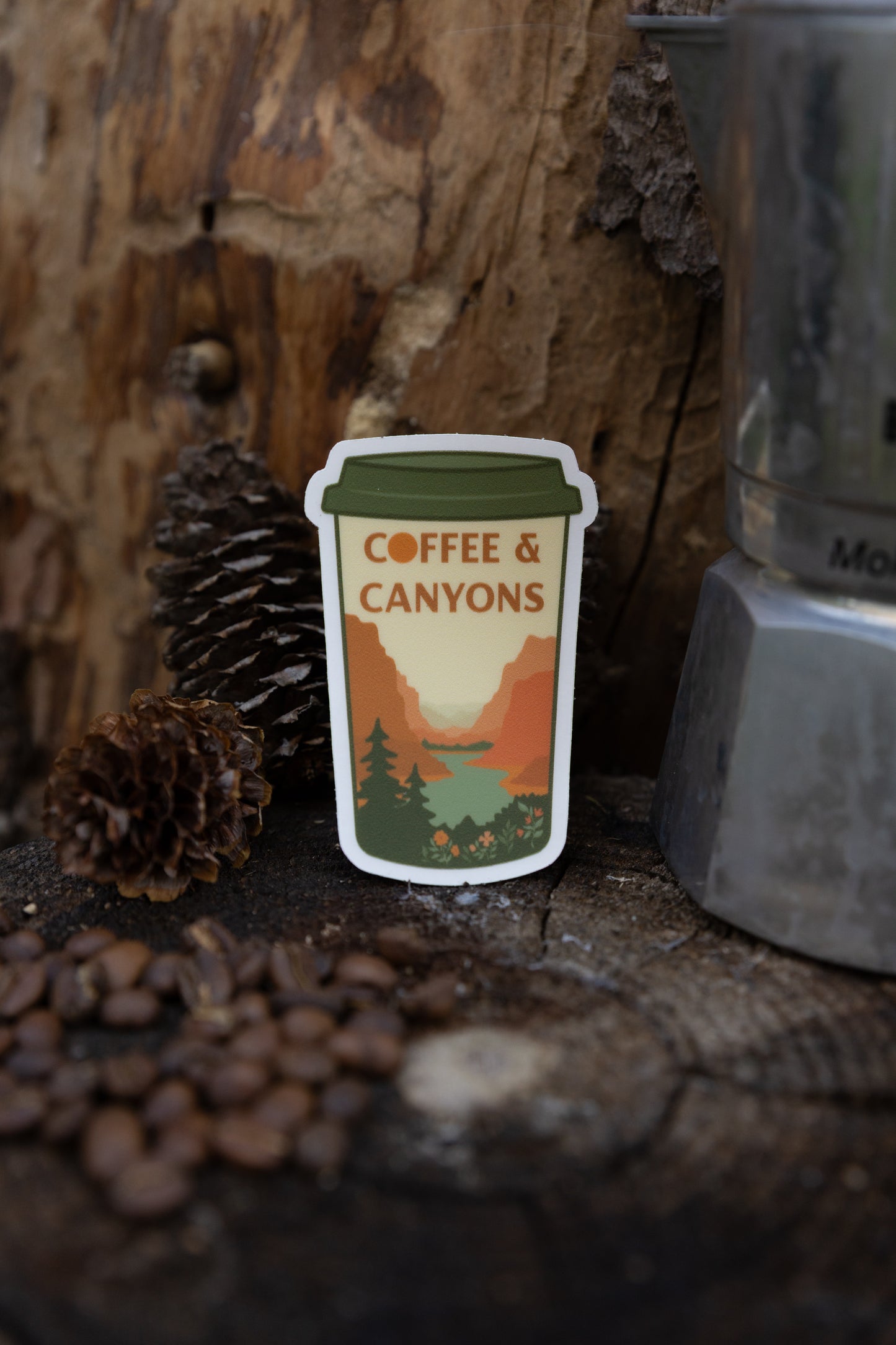 A close-up lifestyle photo of the "Coffee & Canyons" sticker standing upright on a rustic wood stump. The background shows weathered bark, pinecones, and scattered coffee beans, with a silver Moka pot to the right.