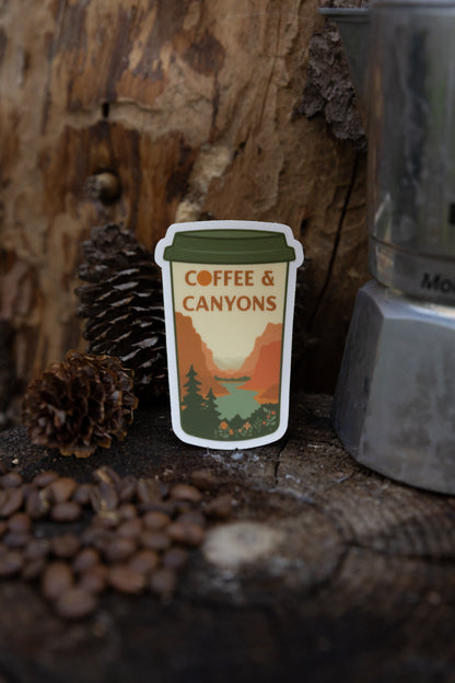 A close-up lifestyle photo of the "Coffee & Canyons" sticker standing upright on a rustic wood stump. The background shows weathered bark, pinecones, and scattered coffee beans, with a silver Moka pot to the right.