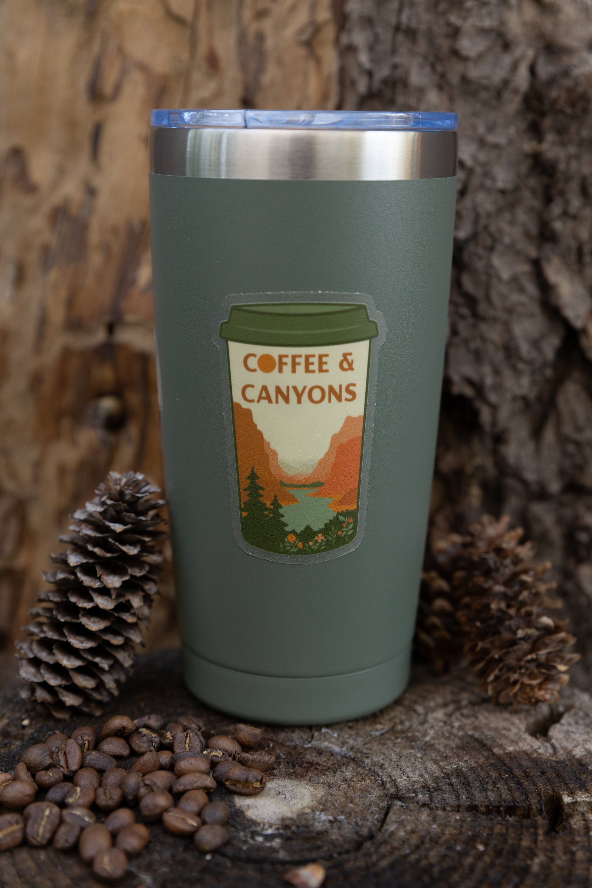 The "Coffee & Canyons" adventure sticker applied to a sage green stainless steel travel tumbler. The tumbler is set against a natural wood and bark background with pinecones and coffee beans at the base.