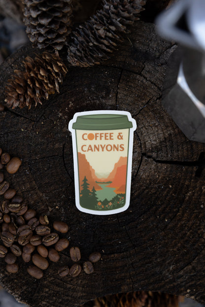 A top-down flat lay view of the "Coffee & Canyons" die-cut vinyl sticker resting on a dark wood tree slice, surrounded by pinecones, coffee beans, and the edge of a Moka pot.