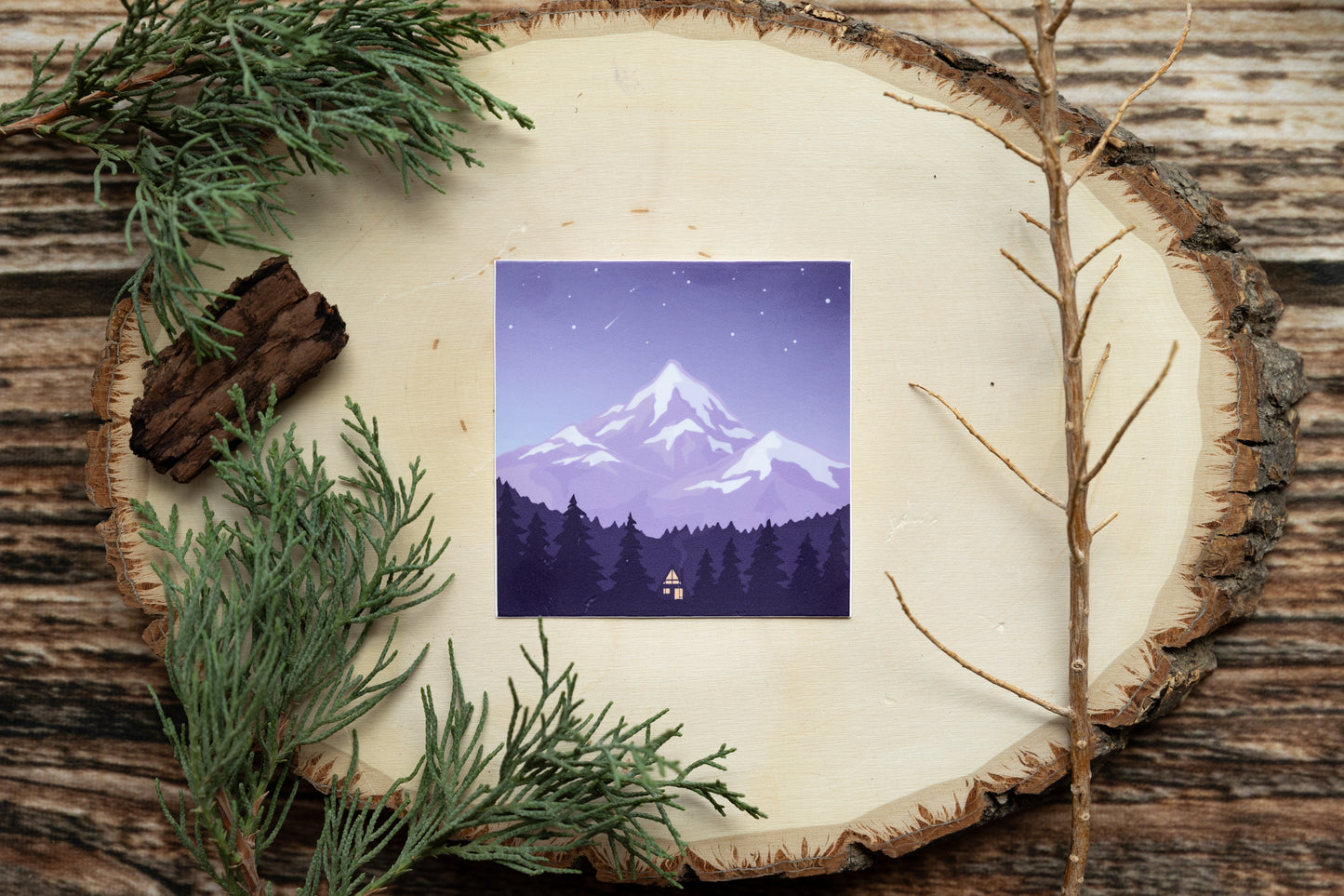 sticker of a small cabin in the woods below a mountain and surrounded by trees